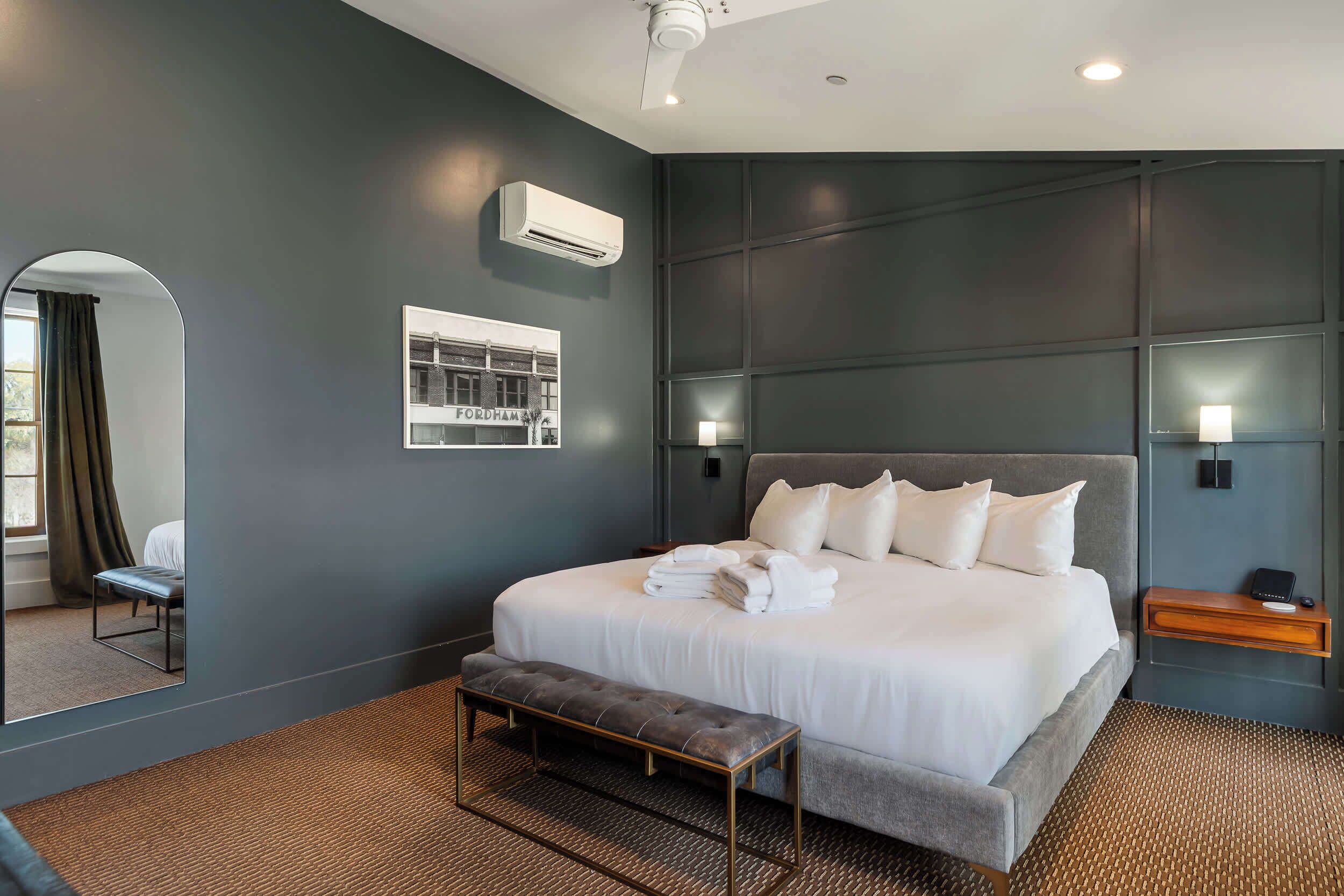 Una camera d'albergo moderna con un letto grande, una panca, uno specchio, un condizionatore d'aria e un quadro incorniciato appeso al muro.