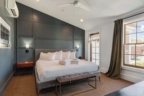 A hotel room with a large bed, a bench, a ceiling fan, an air conditioning unit, a window with curtains, and a view of buildings outside.
