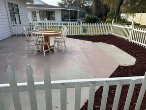 Un patio con cerca blanca de piquetes, una mesa de madera con sillas y una casa al fondo.