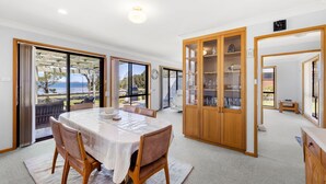 A spacious dining area with a large table, chairs, and a view of the outdoors. There is a wooden cabinet with glass doors and a ceiling light. - The Bay House - Fireplace - Waterfront (Callala Bay)