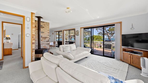 A spacious living room with a large white sofa, a flat-screen TV, and a fireplace. - The Bay House - Fireplace - Waterfront (Callala Bay)
