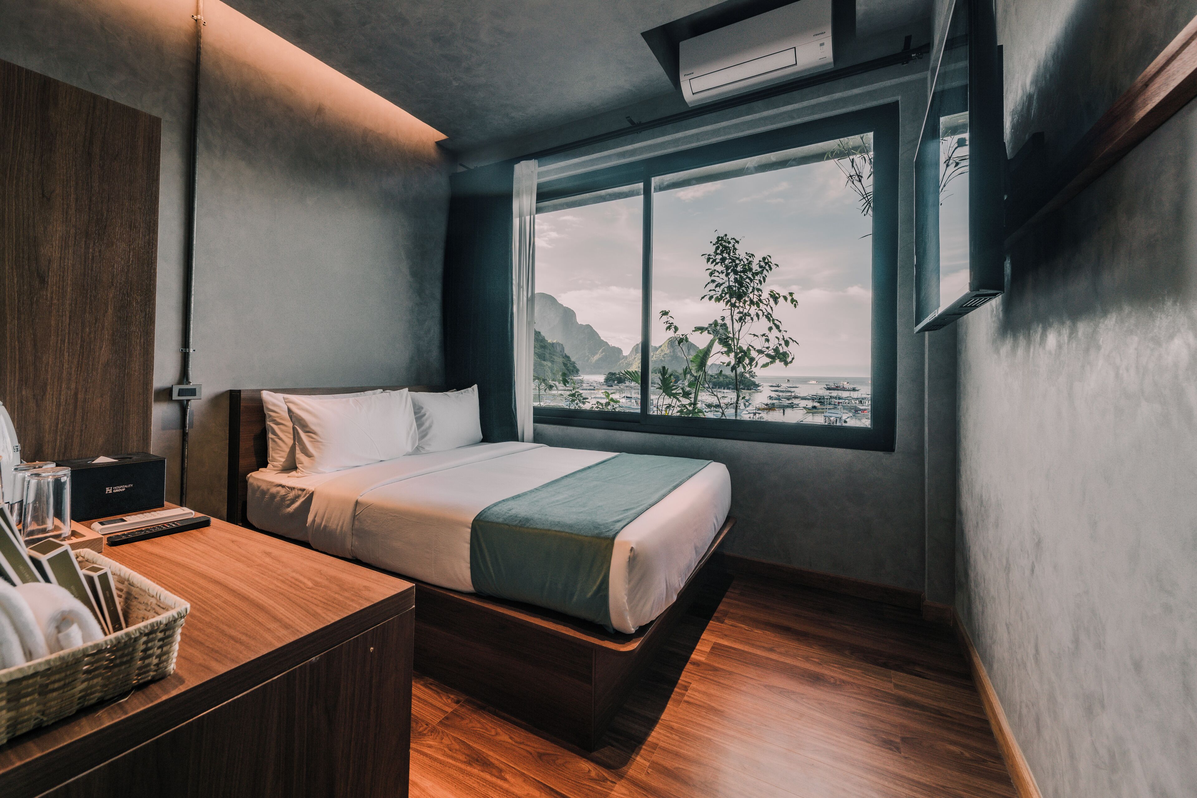 A modern hotel room with a large window, a bed, a wooden desk, and a small table with a basket.