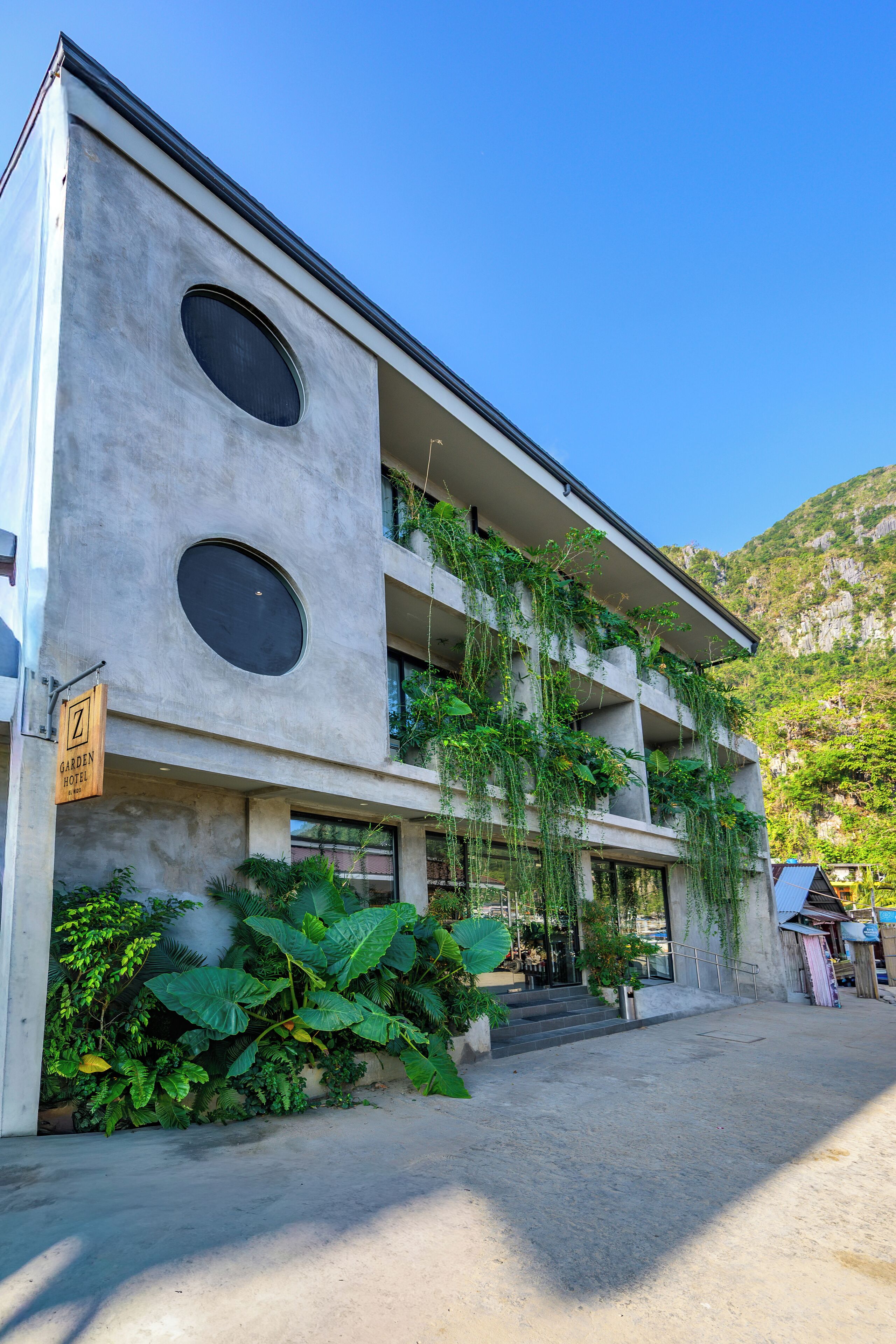 Un hotel moderno con fachada de concreto, amplias ventanas circulares y abundante vegetación.