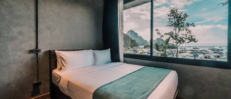 A bedroom with a bed, a window with a view of mountains and boats, and a wooden floor.