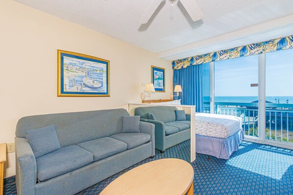 Une chambre d’hôtel avec un canapé, un fauteuil plus petit, un lit, une table et une grande fenêtre donnant sur la mer.