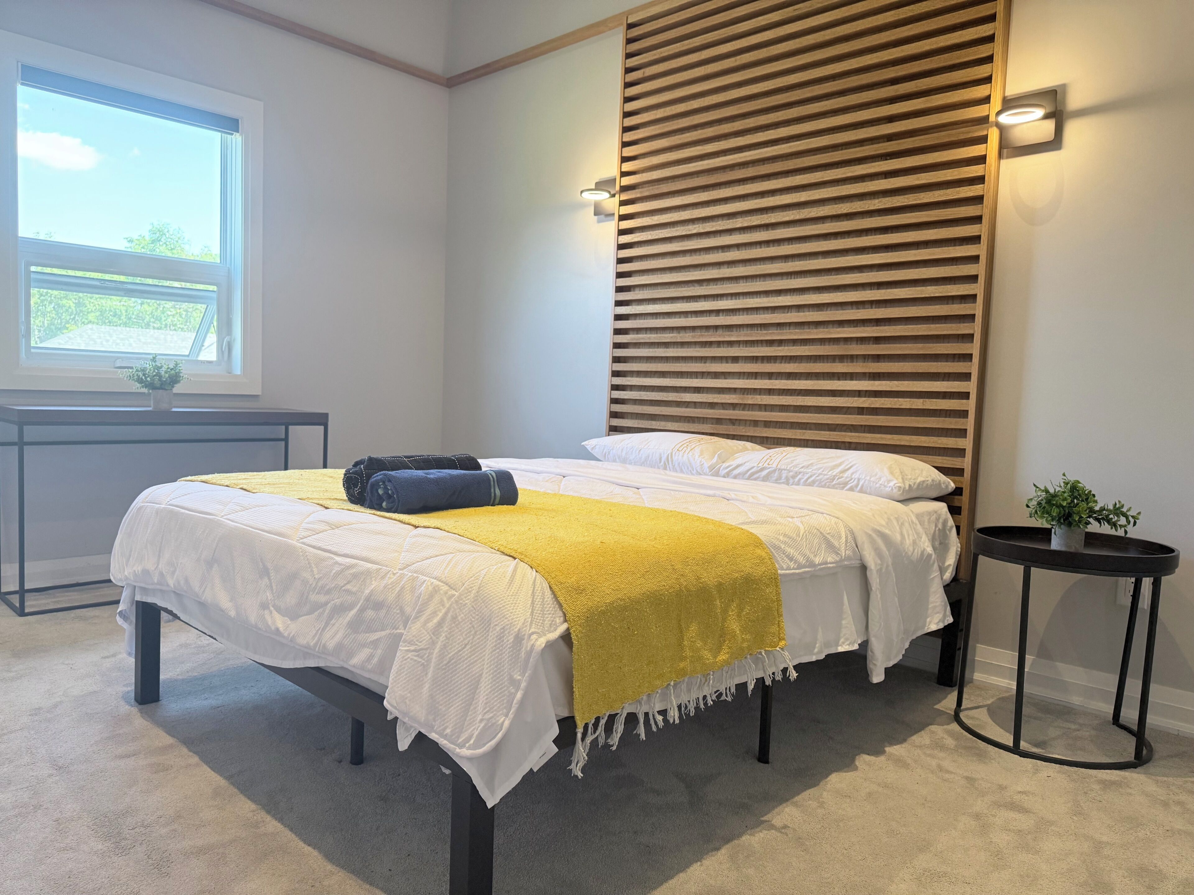 A bedroom with a bed, a bedside table, a nightstand, a window, and a wall-mounted light fixture.