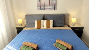 Een keurig opgemaakt bed met een blauw deken, twee opgerolde handdoeken en een schilderij van palmbomen aan de muur.