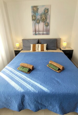 Ein ordentlich gemachtes Bett mit blauer Bettdecke, zwei zusammengerollten Handtüchern und einem Palmenbaum-Gemälde an der Wand.
