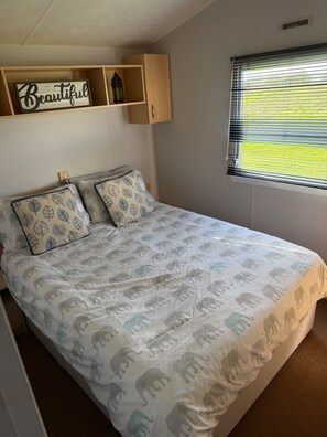 A bedroom with a bed, pillows, a shelf with a sign, and a window with blinds. - Beautiful 3 Bed, Sleeps 6, Wifi, Pet Friendly (Bridlington)