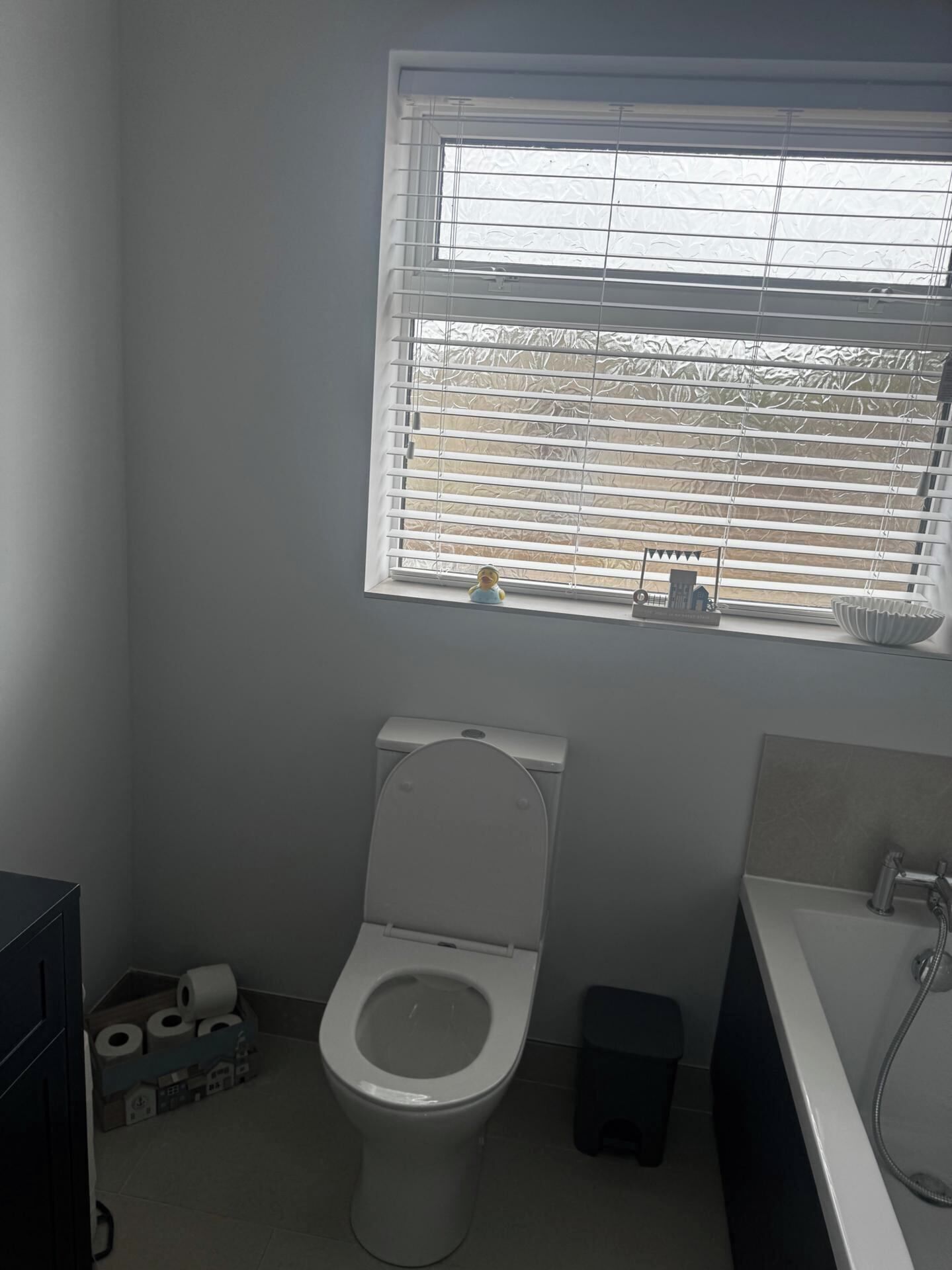 A bathroom with a toilet, a window with blinds, a sink, and a trash bin.