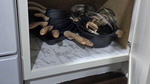 Een keukenkastje met een marmeren aanrechtblad, waarin een zwarte koekenpan, een glazen deksel en houten keukengerei zitten.