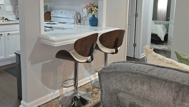 Een moderne keuken met witte kastjes, een magnetron en een bloevas. In de woonkamer staan twee barkrukken en een grijze bank.
