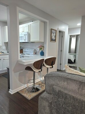 A modern kitchen with white cabinets, a microwave, and a vase of flowers. There are two bar stools and a gray sofa in the living area.