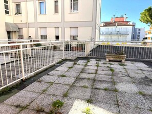 En tagterrasse med gelænder af hvid metal, en stenblomsterkasse og en bygning med vinduer og altaner i baggrunden.
