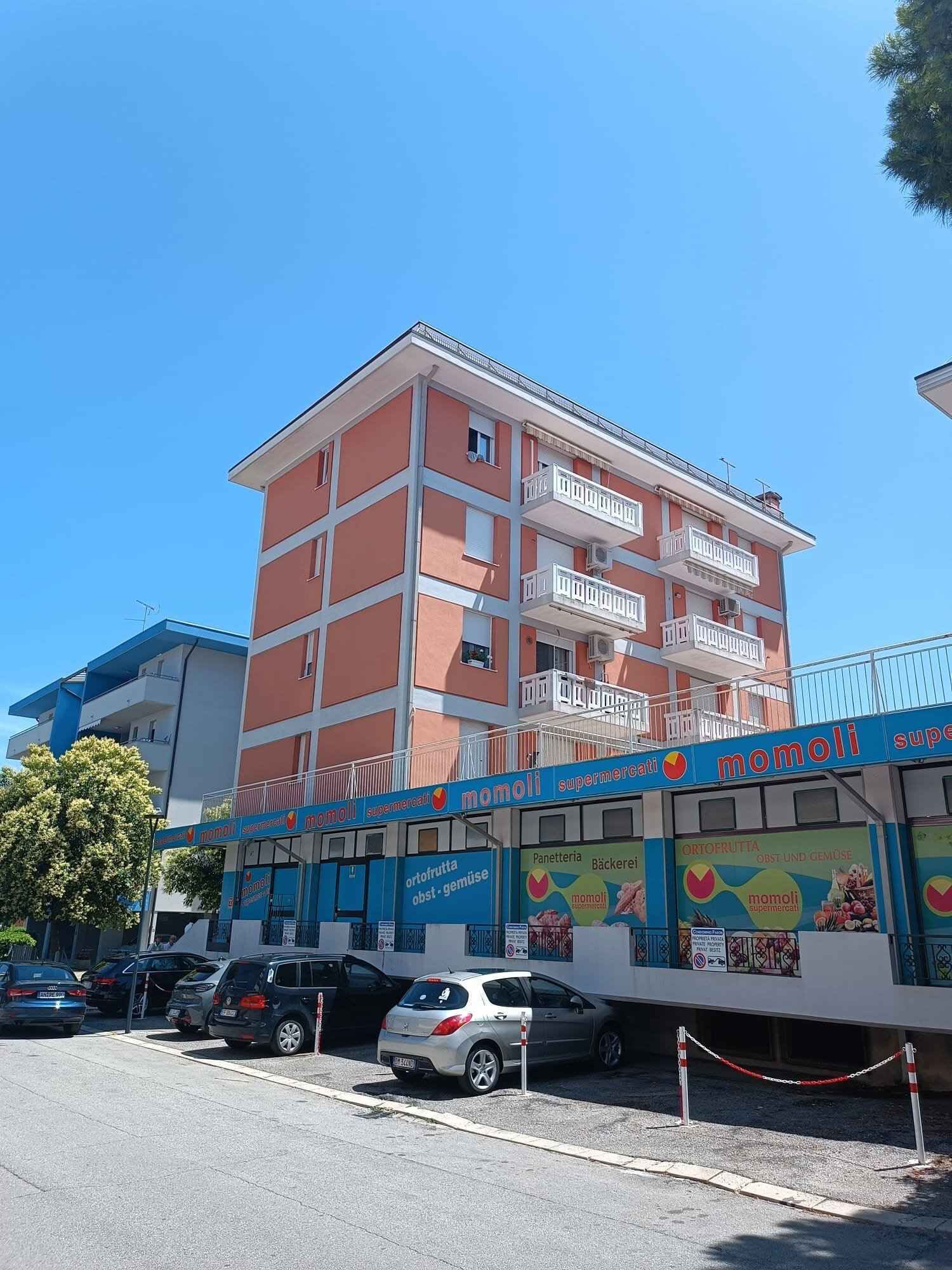A multi-story building with a commercial ground floor featuring a supermarket named 'mobili' and a parking area with several cars.