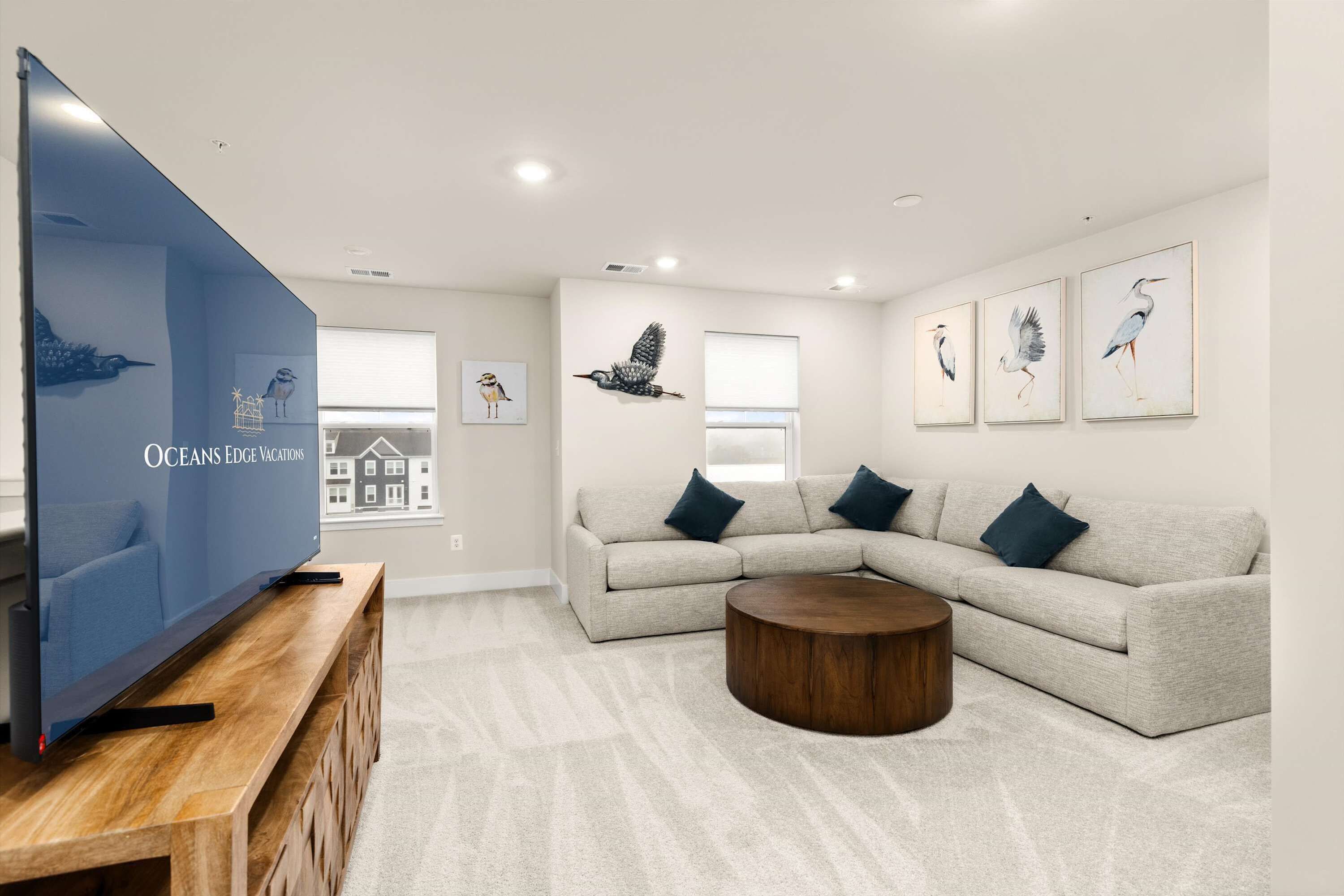A modern living room with a large flat-screen TV, a grey sectional sofa, a round wooden coffee table, and wall-mounted artwork.