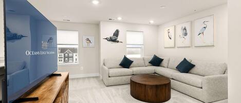 A modern living room with a large flat-screen TV, a grey sectional sofa, a round wooden coffee table, and wall-mounted artwork.