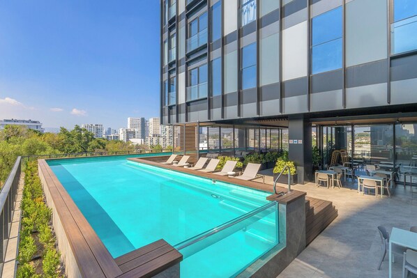 A rooftop pool with lounge chairs and a dining area, surrounded by a modern building with large windows.