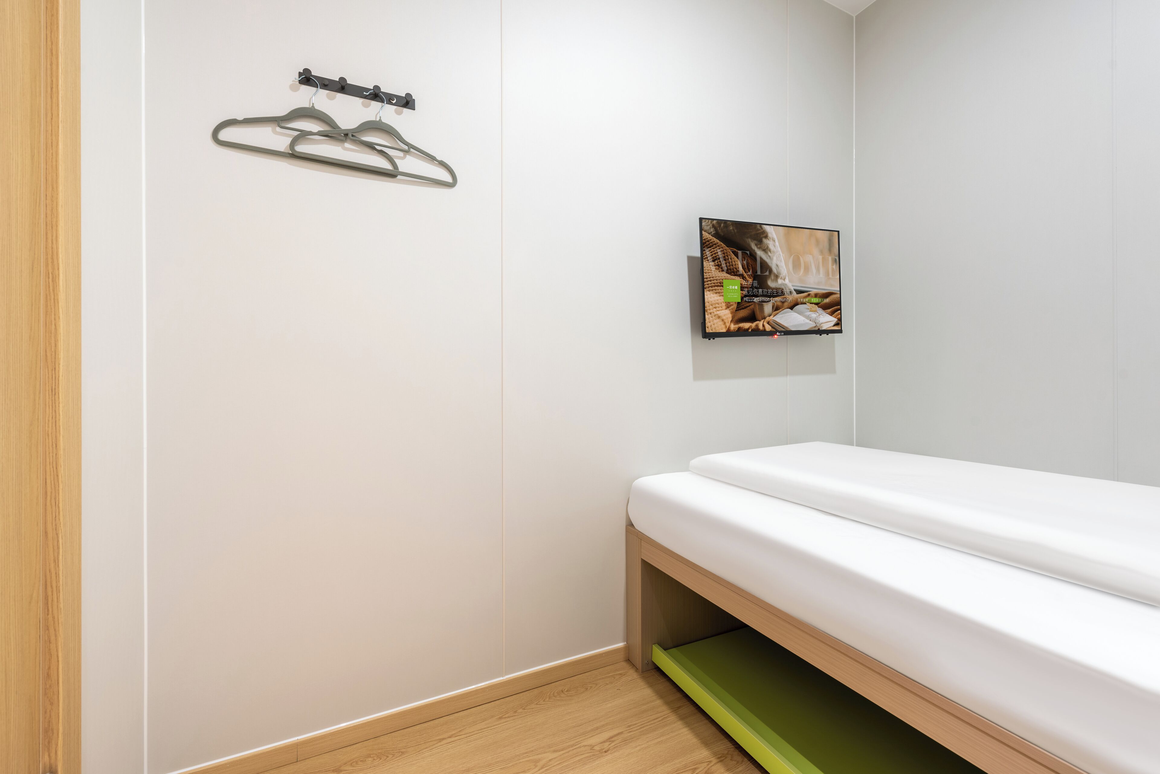 A minimalist room with a wooden floor, a white bed, a green shelf, and a wall-mounted picture.
