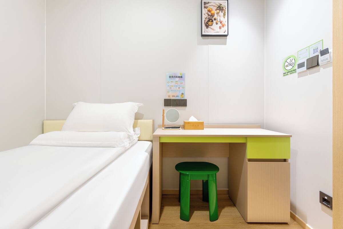 Une petite pièce, bien rangée, avec un lit, un bureau agrémenté d’un tabouret vert, une station de recharge fixée au mur et un tableau encadré.