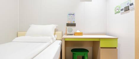 A compact room with a bed, a desk with a green stool, a wall-mounted charging station, and a framed picture.