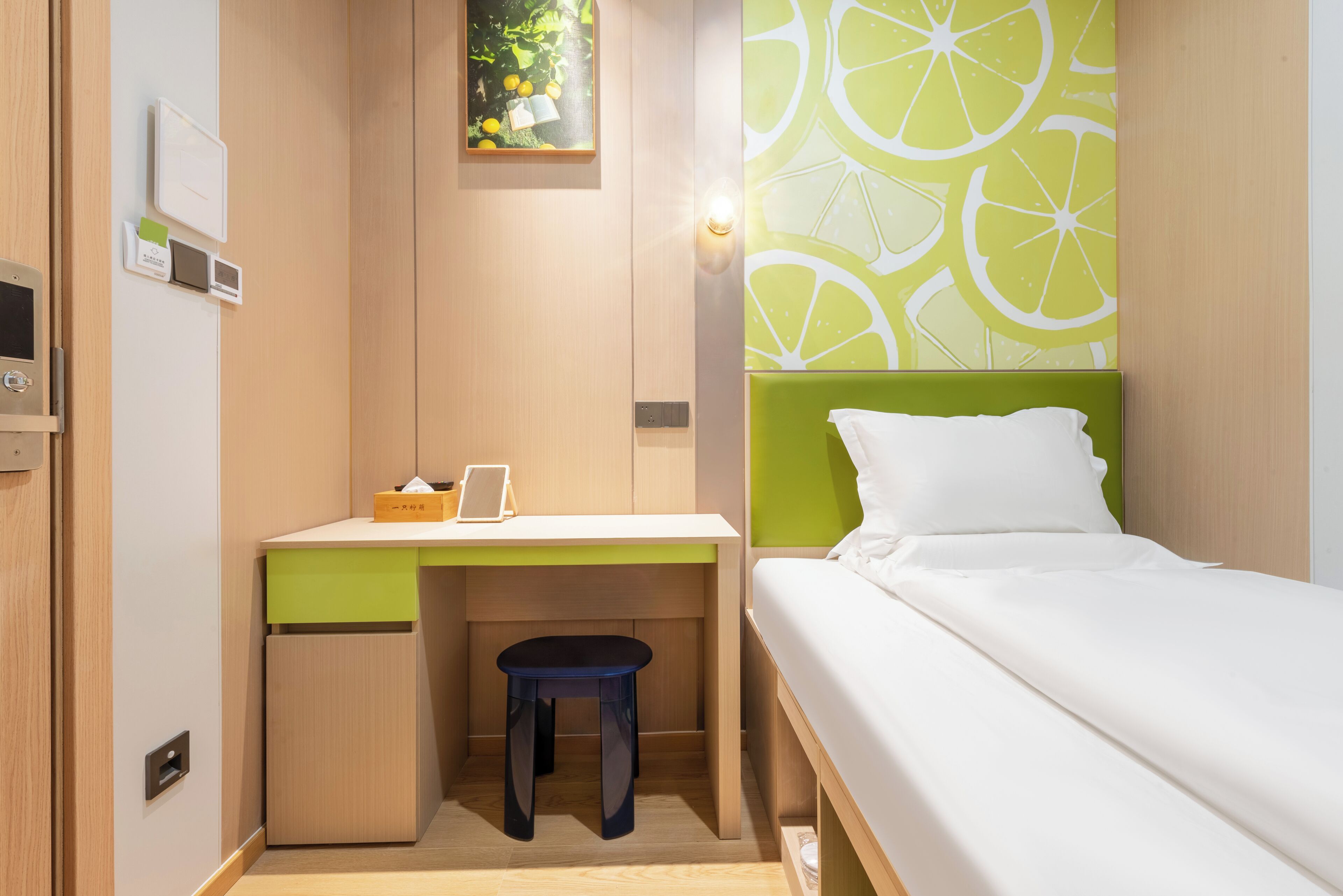 A compact room with a bed, a desk, and a stool. There is a wall-mounted light and a small window.