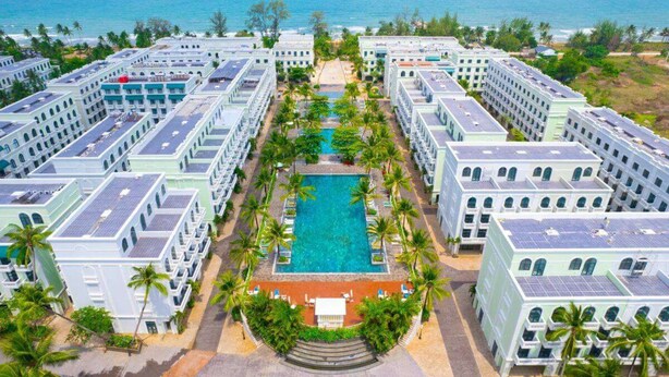 Un resort con varias edificaciones, una piscina central y palmeras.