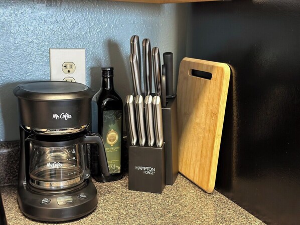 Un comptoir de cuisine avec une machine à café de la marque Mr. Coffee, une bouteille d’huile d’olive, un bloc à couteaux Hampton Forge et une planche à découper en bois.