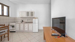 Cocina compacta con alacenas blancas, un refrigerador, estufa y pileta. Hay un banco de madera y un televisor sobre un mueble bajo.