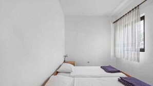 Una habitación pequeña y limpia con dos camas individuales, una mesita de noche, una lámpara y una ventana con cortinas translúcidas.