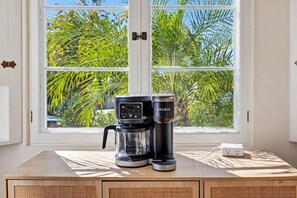 Eine Kaffeemaschine von Keurig und eine Kaffeekanne auf einem Holztisch neben einem Fenster mit Blick auf Palmen.