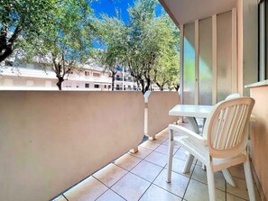 A balcony with a table and chairs, a window with blinds, and a view of buildings and trees. - Holiday apartment with air conditioning and balcony (Bibione)
