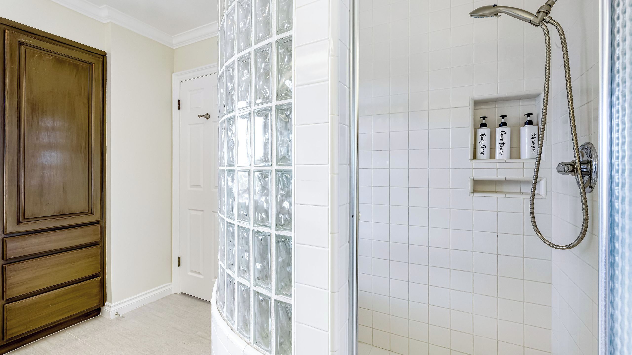 Een badkamer met witte betegelde wanden, een douche met een handdouche en een douchewand van glasblokken.