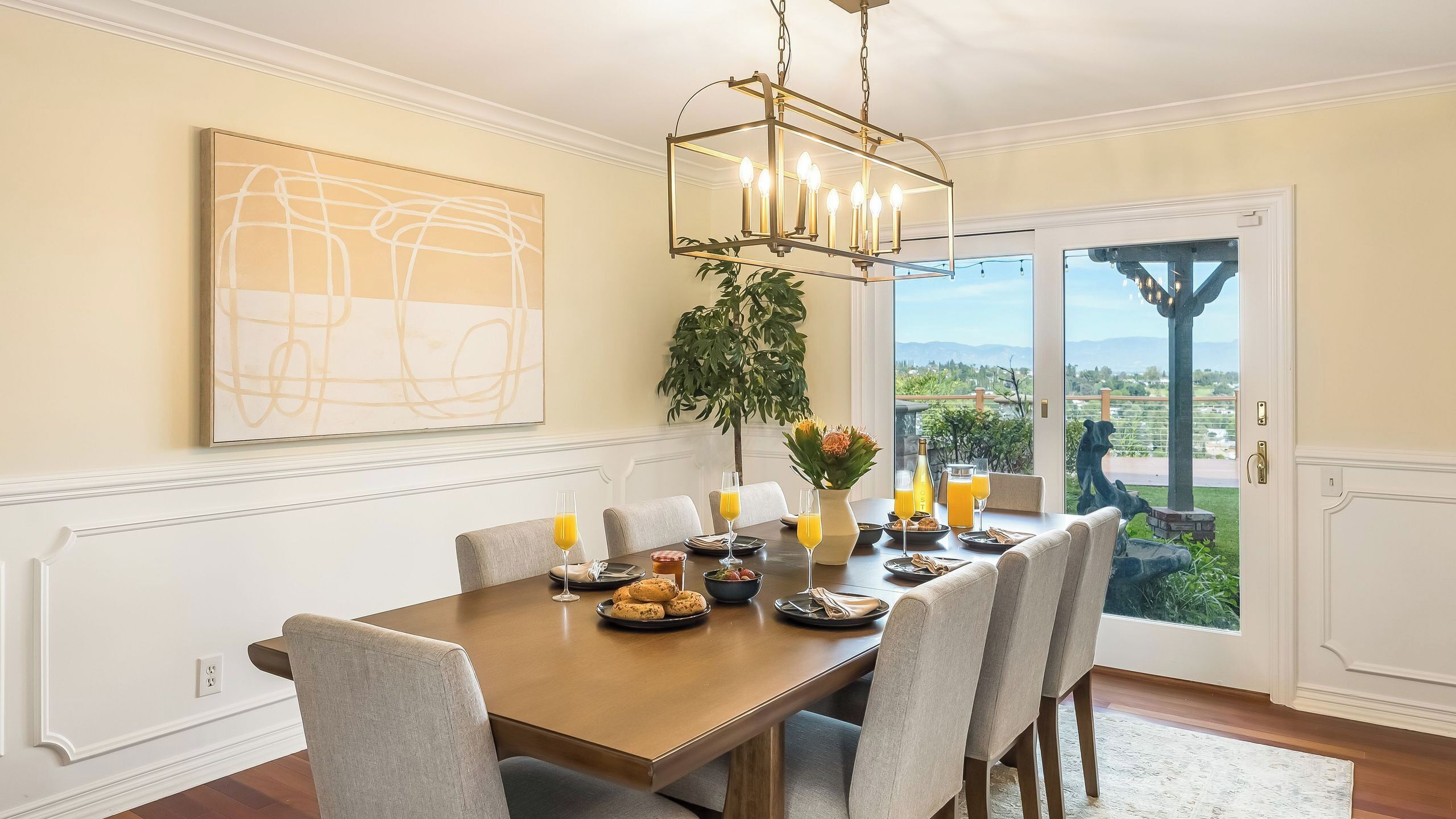 Uma sala de jantar com mesa de madeira, cadeiras, um lustre, um quadro abstrato grande e vista pela porta de vidro.