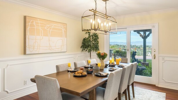 A dining area with a wooden table, chairs, a chandelier, a large abstract painting, and a view through a glass door. - Twilight Terrace by Avantstay West Hills Retreat Hot Tub, BBQ + EV Charger, Near Malibu (West Hills)