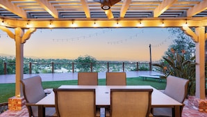 An outdoor patio with a table and chairs, string lights, and a view of the cityscape. - Twilight Terrace by Avantstay West Hills Retreat Hot Tub, BBQ + EV Charger, Near Malibu (West Hills)