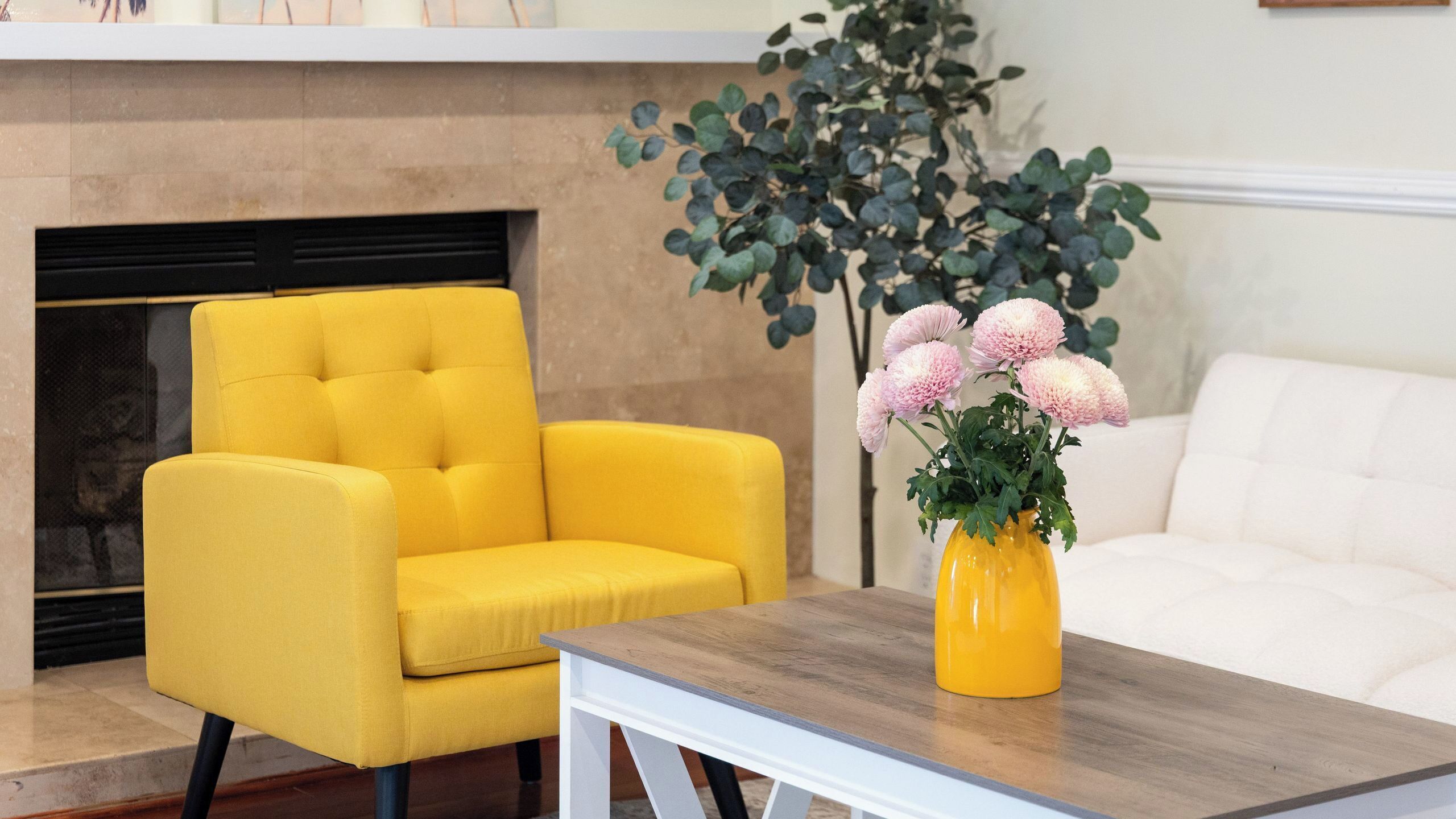 A yellow armchair, a white sofa, and a wooden table with a vase of flowers in a yellow vase.