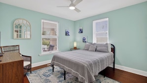 Une chambre à coucher comprenant un lit, un bureau, une chaise, deux fenêtres avec stores, un ventilateur de plafond et des œuvres d’art accrochées aux murs.