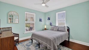 Quarto com cama, escrivaninha, cadeira, duas janelas com persianas, ventilador de teto e obras de arte nas paredes.