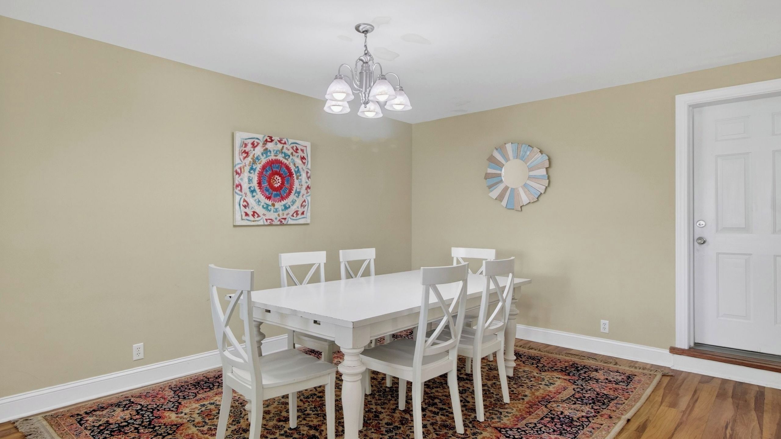 A dining room with a white table and chairs, a chandelier, a patterned rug, and two wall decorations.