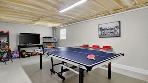 Une table de ping-pong, quatre chaises rouges, un téléviseur et des étagères remplies de jouets et de jeux, le tout dans une pièce aux murs blancs et au plafond en bois.