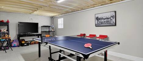 A ping pong table with four red chairs, a TV, and shelves with toys and games in a room with white walls and a wooden ceiling.