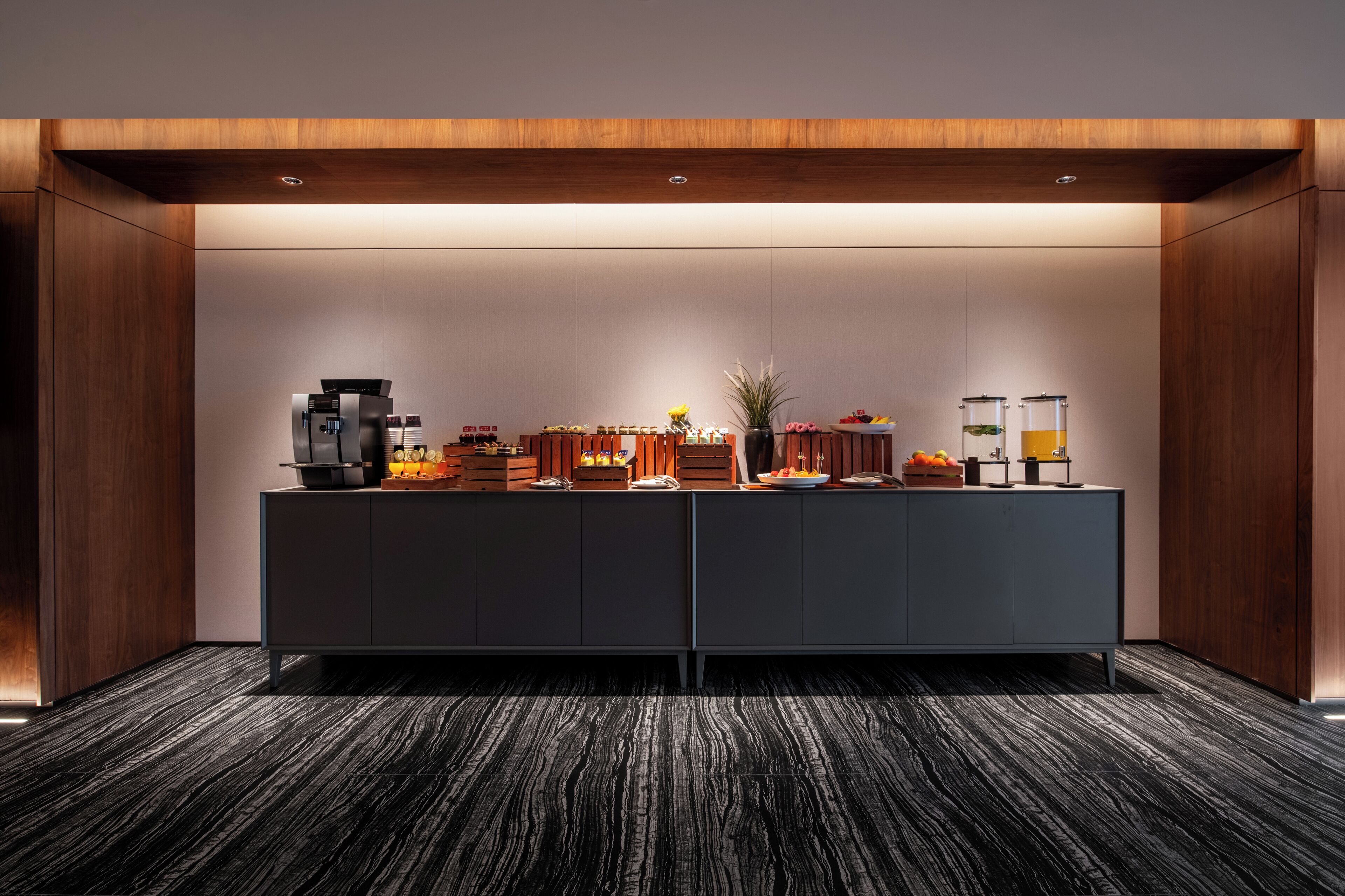 A modern hotel reception area with a dark wood panel design, a central counter with various amenities, and a carpeted floor.