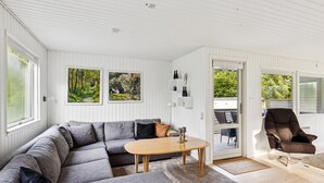 Ett modernt vardagsrum med en hörnsoffa, ett soffbord i trä och en brun fåtölj. På väggen hänger två tavlor, taket är vitt och det finns en dörr som leder ut.