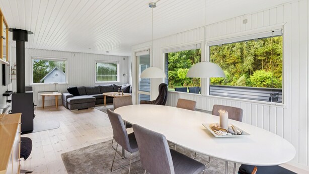 Ett modernt vardagsrum med ett stort ovalt bord, grå soffor och utsikt över grönskan genom stora fönster.