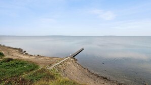 En kustvy med en träbrygga som sträcker sig ut i stilla vatten, omgiven av en stenig strand och gles vegetation.