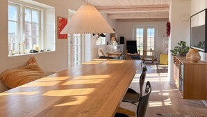 A spacious dining area with a long wooden table, a hanging light fixture, and large windows allowing natural light. - 2 bedroom nice home in Fanø (Fanø)