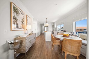 A modern kitchen with a central island, white cabinetry, and a dining area with wicker chairs. - The Boardwalk - Oceanview Unit on Bulcock Beach (Caloundra Region)