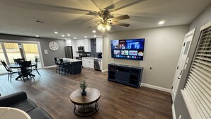 Un vaste espace de vie doté d’un ventilateur de plafond, d’un téléviseur fixé au mur et d’un coin repas avec des chaises et une table.