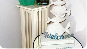 A teal vase with a plant, a glass container with rocks and plants, and a white fish-shaped decoration on a white console table.